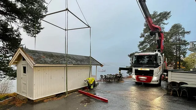 Hytte løftes med kran ved regnvåt parkeringsplass.