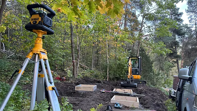 Byggeplass med minigraver og laserutstyr i skog.